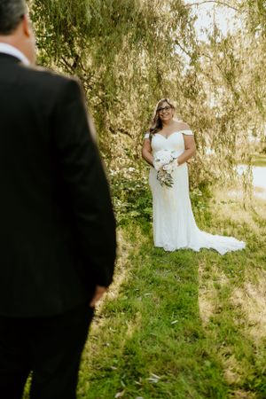 A bride in a white lace gown stands on a grassy hill under weeping willow trees.