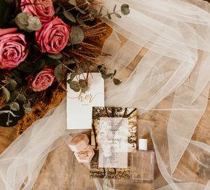 Wedding details flat lay with pink roses, tulle veil, and gold-framed invitation on rustic surface.