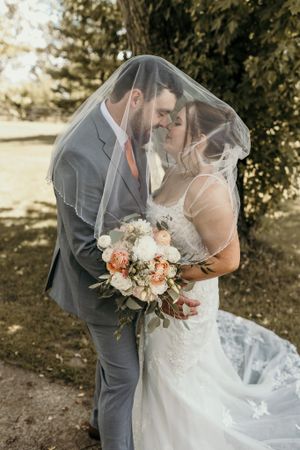 Wedding couple sharing intimate moment under veil with peach and white bouquet.
