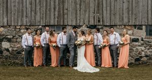 Wedding party poses in front of rustic barn with bridesmaids in peach dresses and groomsmen in gray suits.