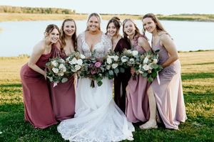 Wedding party in mauve bridesmaid dresses pose together holding bouquets in an outdoor field setting.