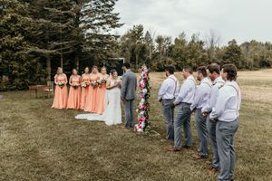 Wedding ceremony setup with bridal party in peach and gray attire standing in outdoor field.