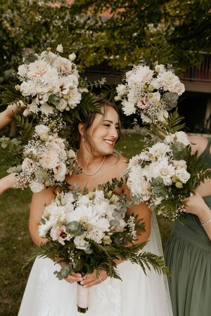 Elegant wedding party in sage green dresses and white floral bouquets pose together in a garden setting.