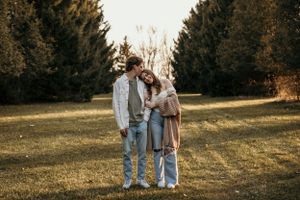 Couple stands close together in front of evergreen trees during golden hour in a field.