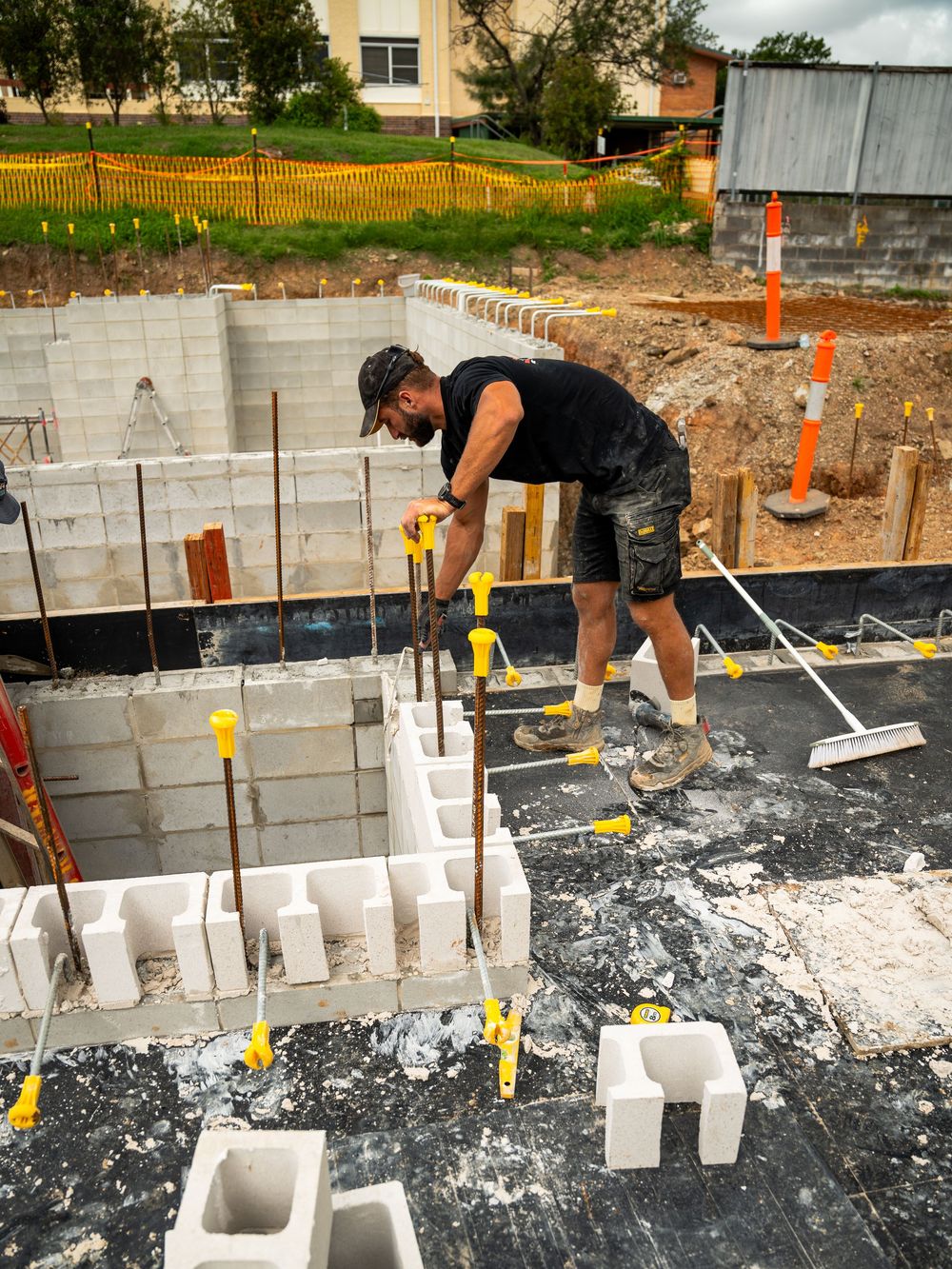 Construction worker laying concrete blocks for a foundation wall with yellow level markers visible on site.