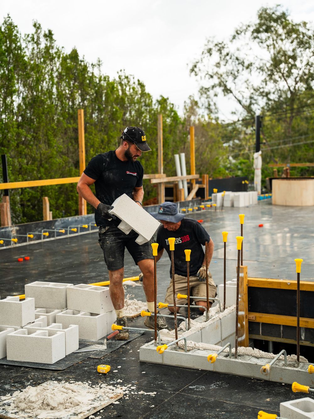 Construction workers assembling white concrete blocks on a raised platform with yellow safety markers and wooden supports.