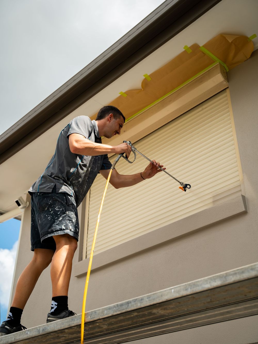A person in shorts and a t-shirt uses a pole-mounted tool to clean the exterior of a house near a window.