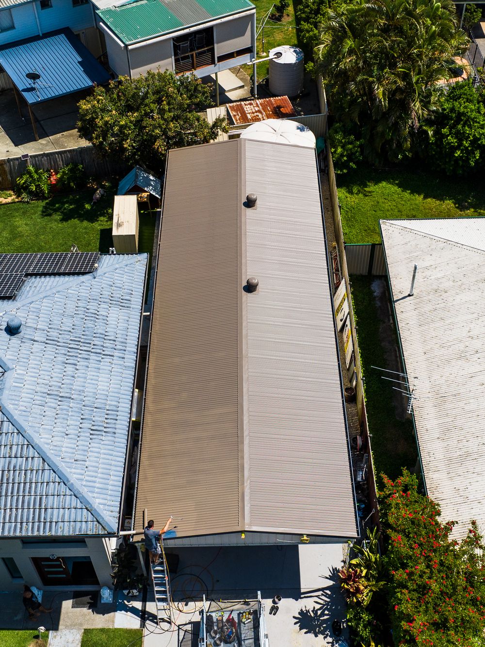 Aerial view of residential rooftops and solar panels with lush green trees surrounding the houses on a sunny day.