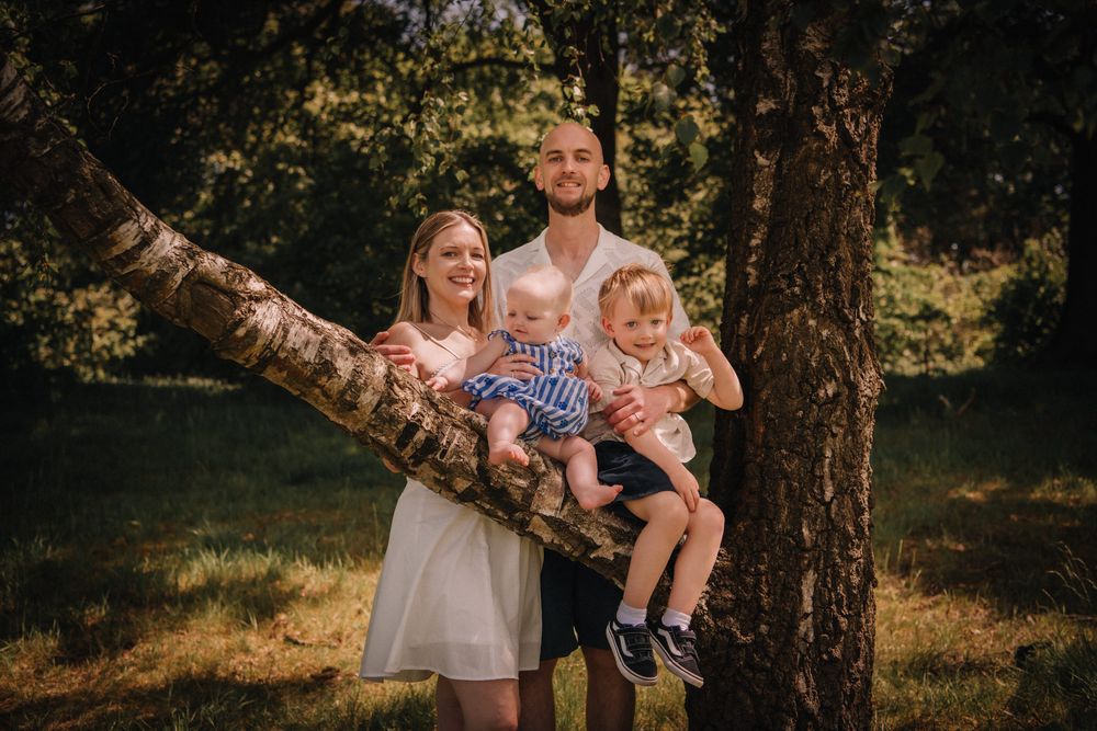Happy family of four posing outdoors near a tree in a sunny forest, parents holding two young children.