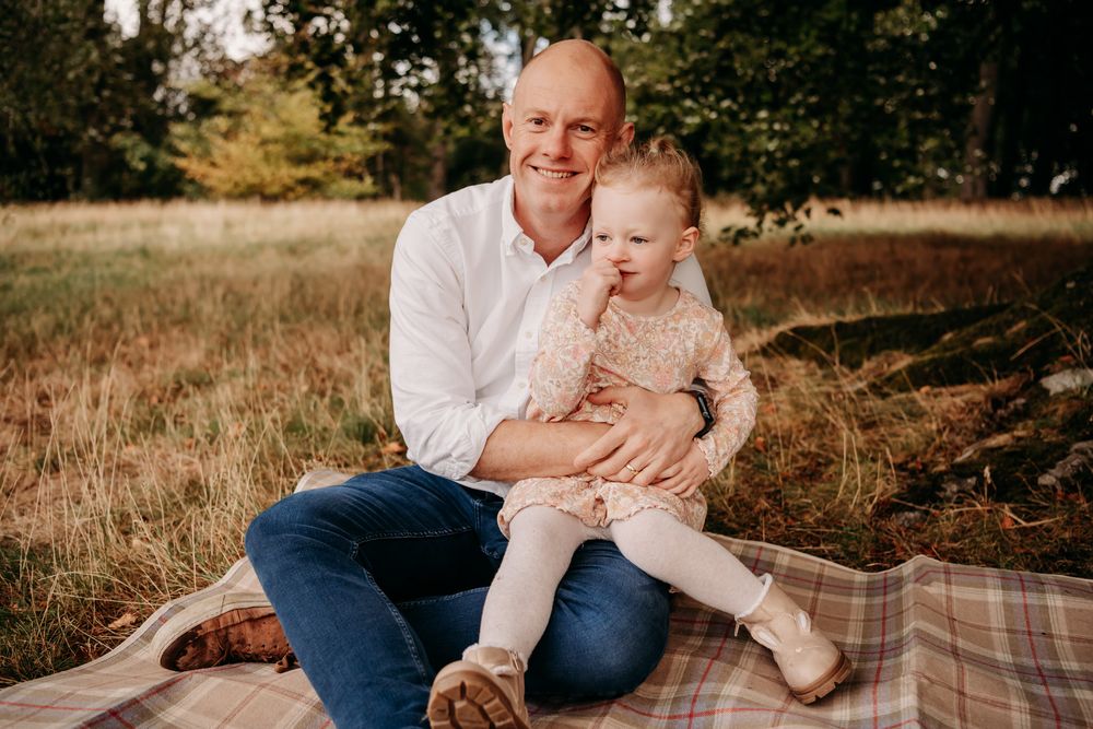 Candid outdoor family portrait with everyone sitting on plaid blanket under tree branches.