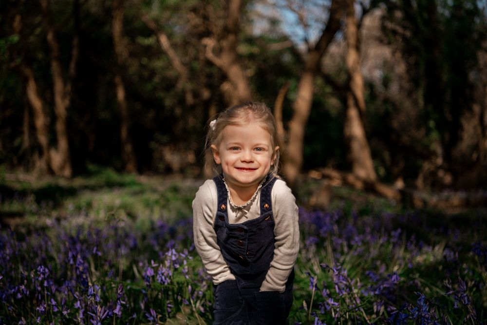 Young girl smiling in a field of purple bluebells surrounded by trees on a sunny day.
