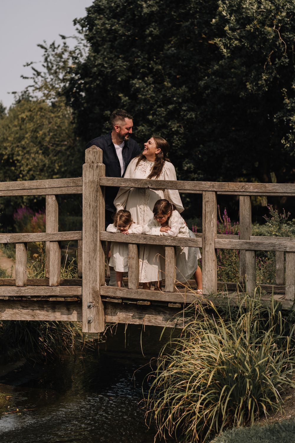 A family of four poses on a rustic wooden bridge surrounded by lush greenery, with parents sharing a loving glance.