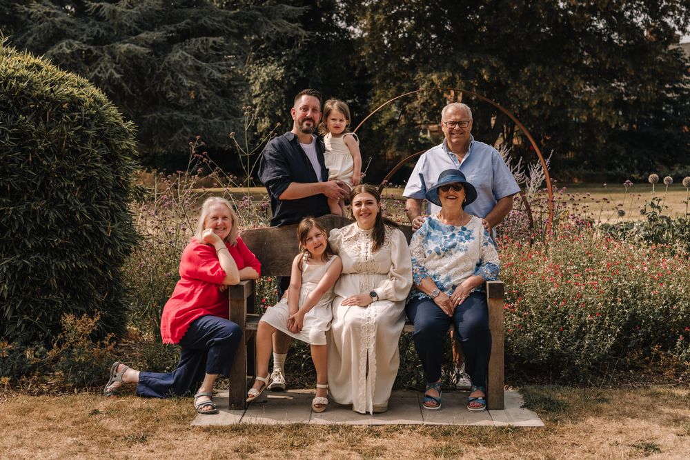 Multi-generational family portrait posing together on a garden bench surrounded by colorful flowers and lush greenery.