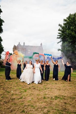 LGBT wedding couple and bridal party holding smoke bombs in the colours of the pride flag with hotel in background, Dundee Scotland