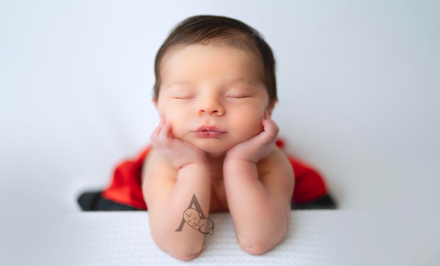 Sleeping baby in red outfit resting head on hands against white background.