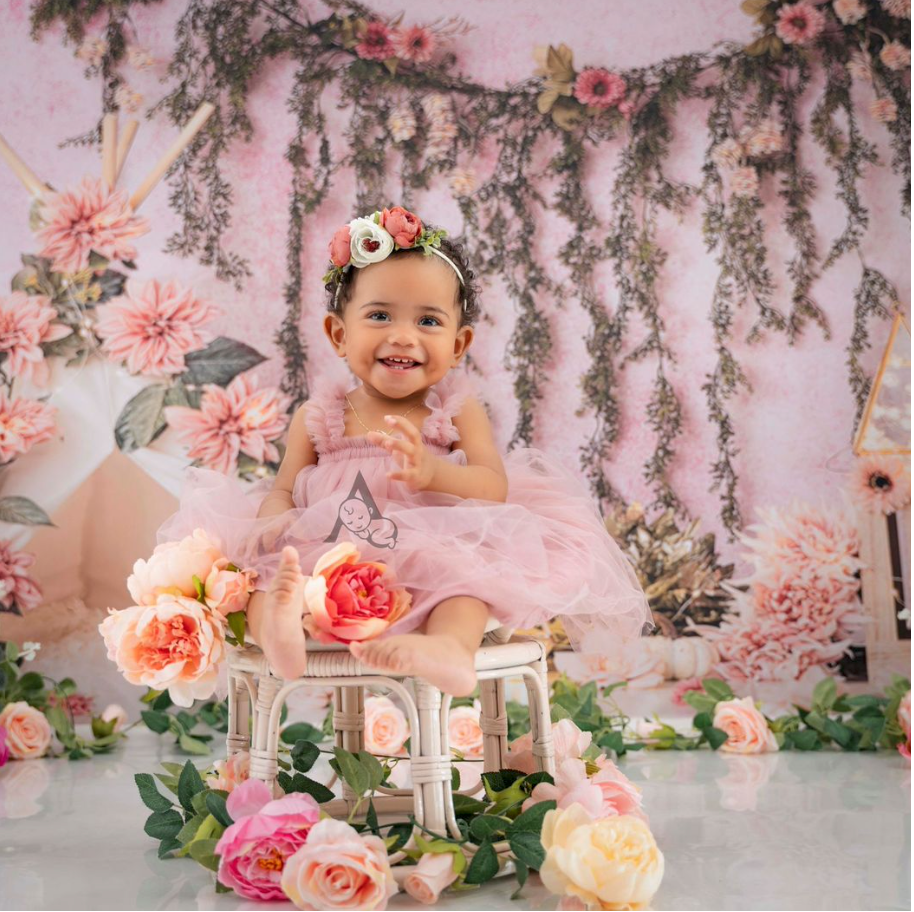 Happy baby in pink tulle dress sitting among roses and floral decorations.