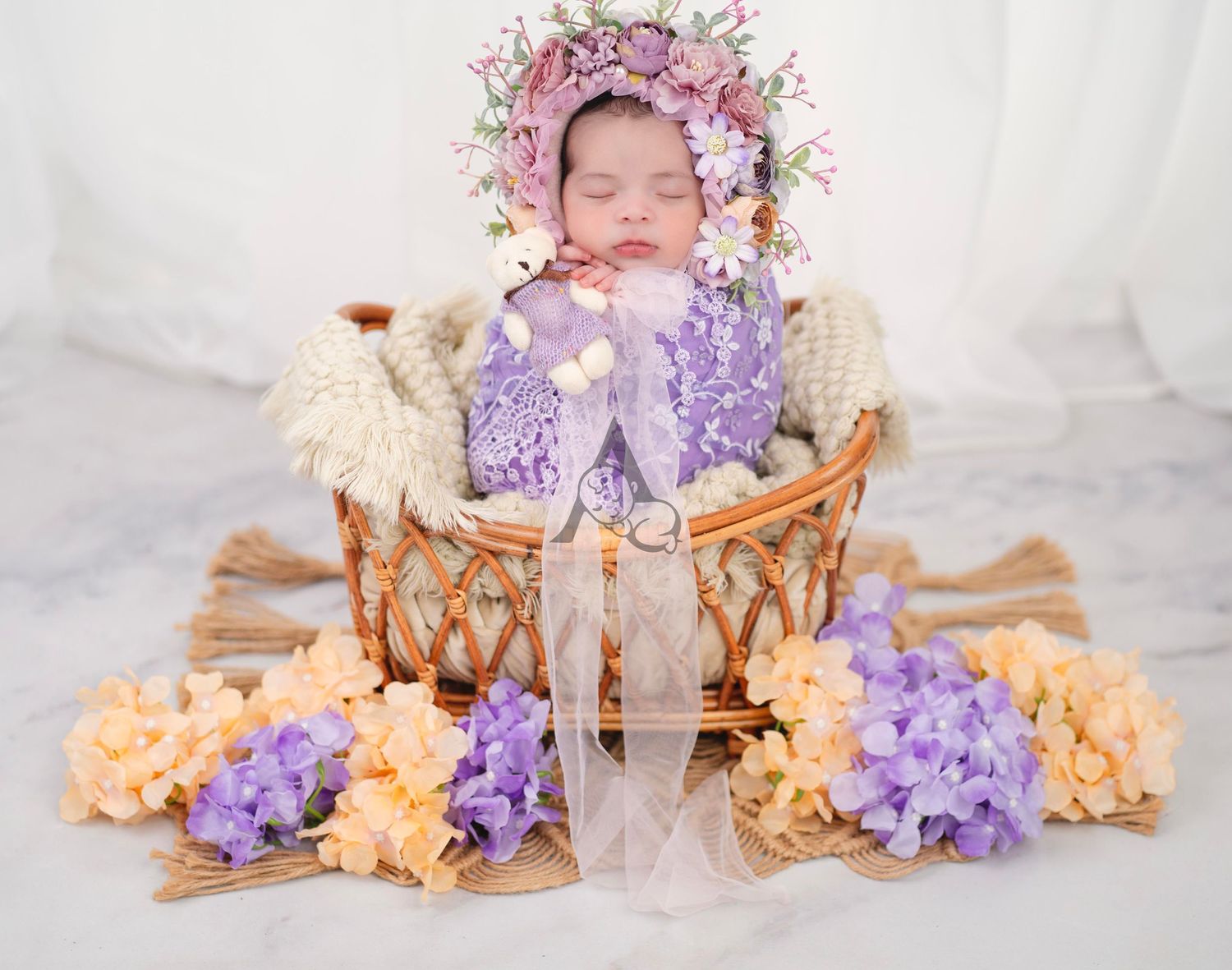 Baby sleeping in wicker basket surrounded by purple and peach flowers with floral crown.