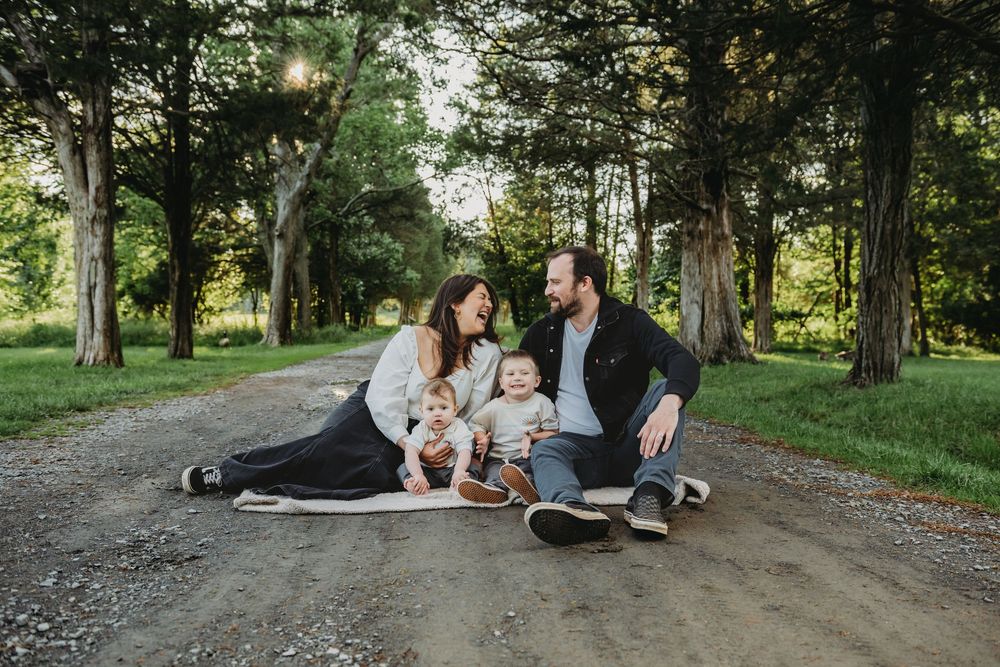 A family with a toddler sits together on a blanket on a tree-lined dirt path in a peaceful summer setting.