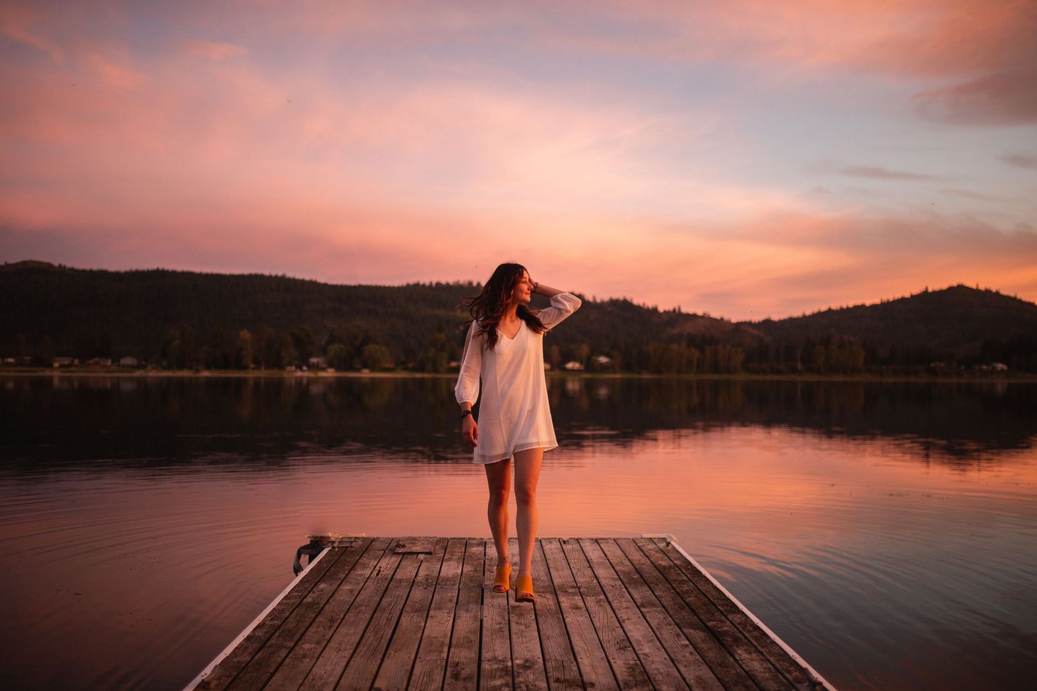 Woman in white dress standing on wooden dock at sunset with pink sky reflecting on calm lake.