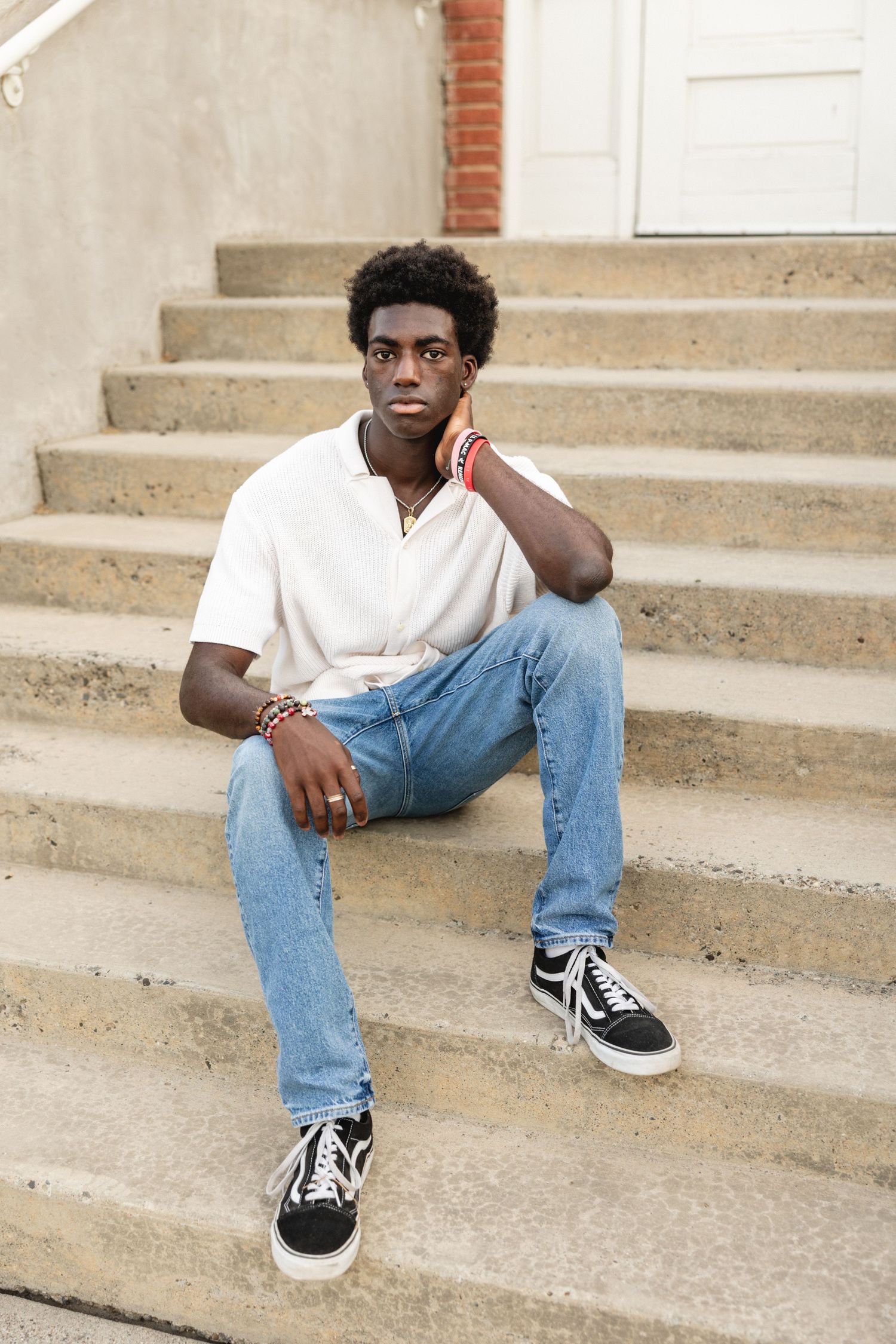 Young Black man in white polo, jeans, and Vans sneakers sitting on concrete steps outdoors.
