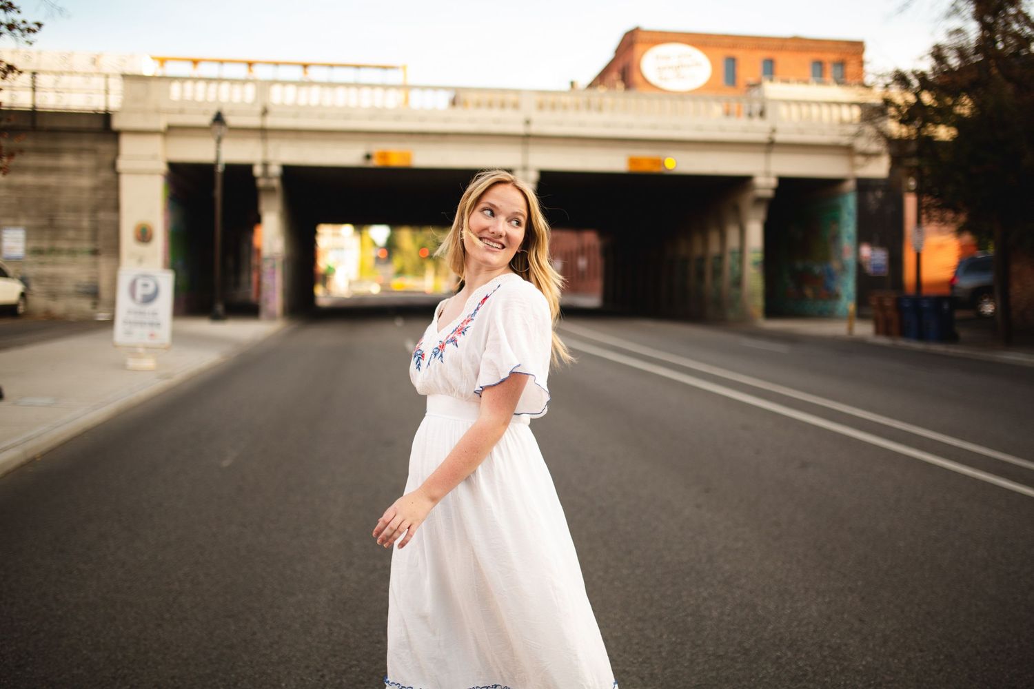 Smiling blonde woman in white midi dress standing on urban road with graffiti bridge backdrop.