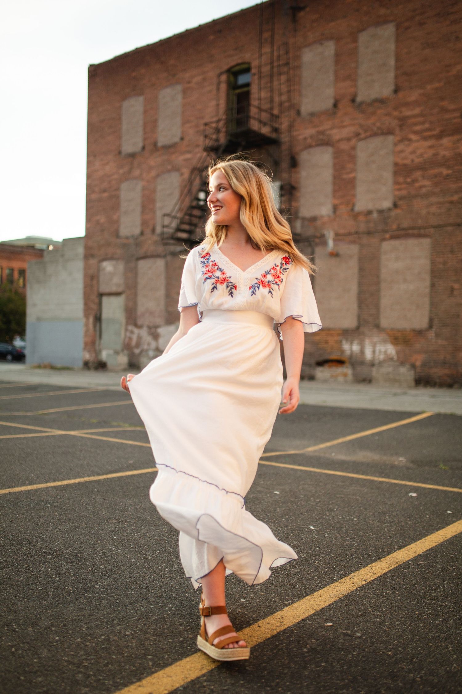 Woman in white floral embroidered maxi dress twirling in urban parking lot with brick building backdrop.