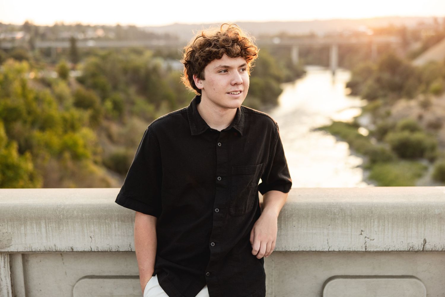 Teen boy with curly hair in black shirt leaning on bridge railing with river and trees at golden hour.