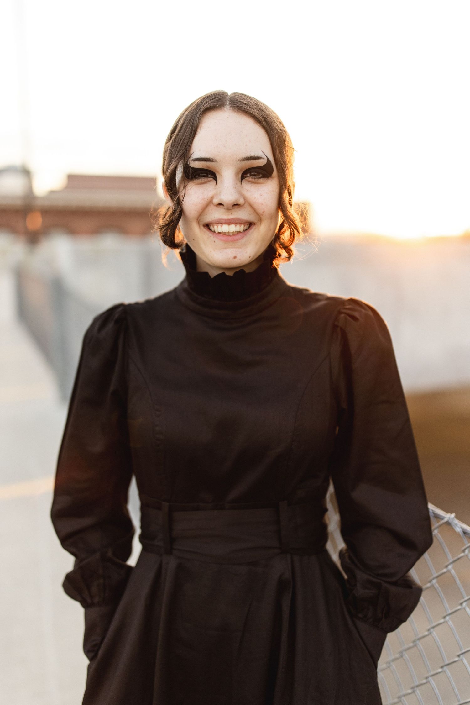 Smiling young woman in black turtleneck dress with dramatic eye makeup, posing outdoors at golden hour sunset.