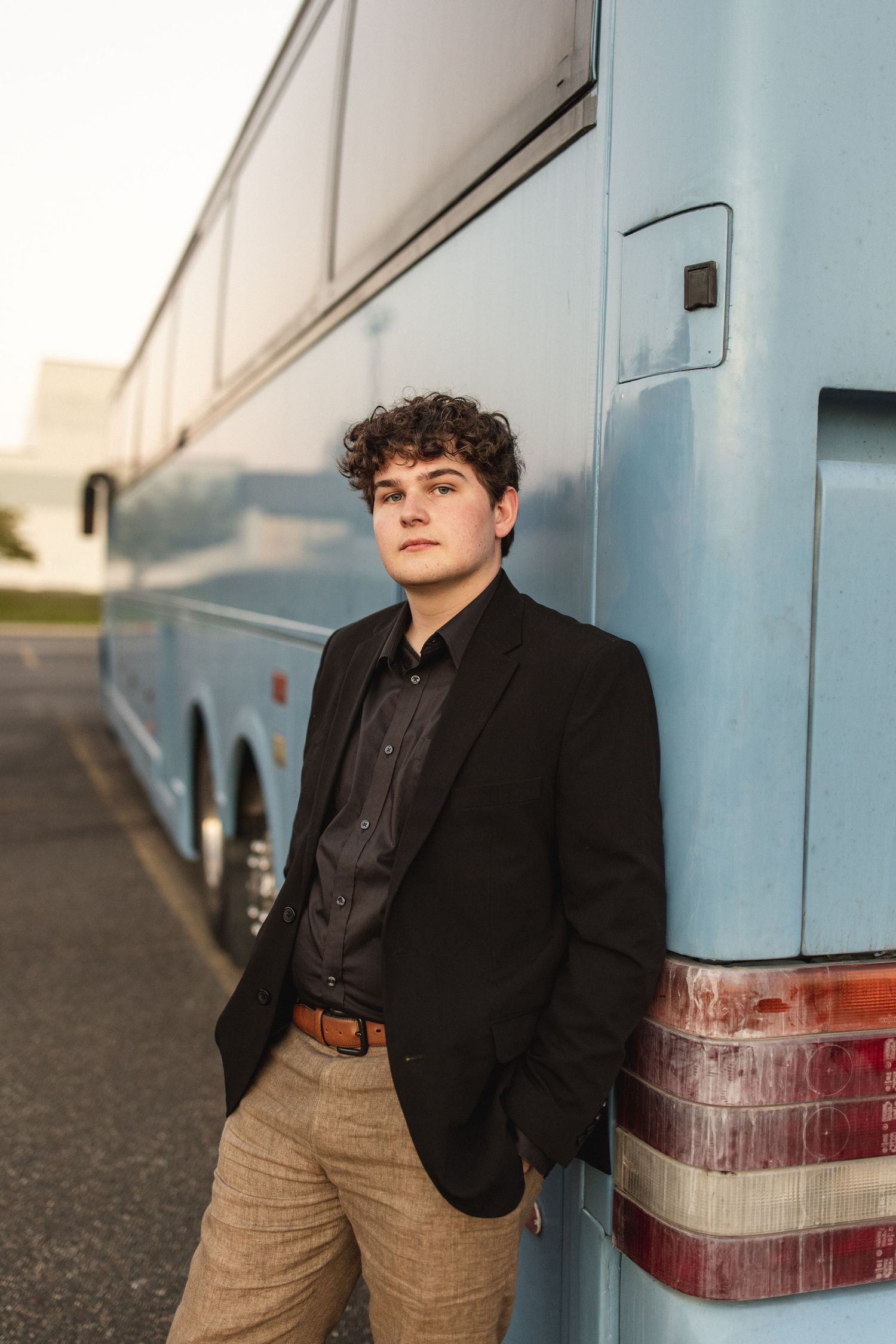 Young man in black blazer and tan pants leaning against a blue bus outdoors at golden hour.