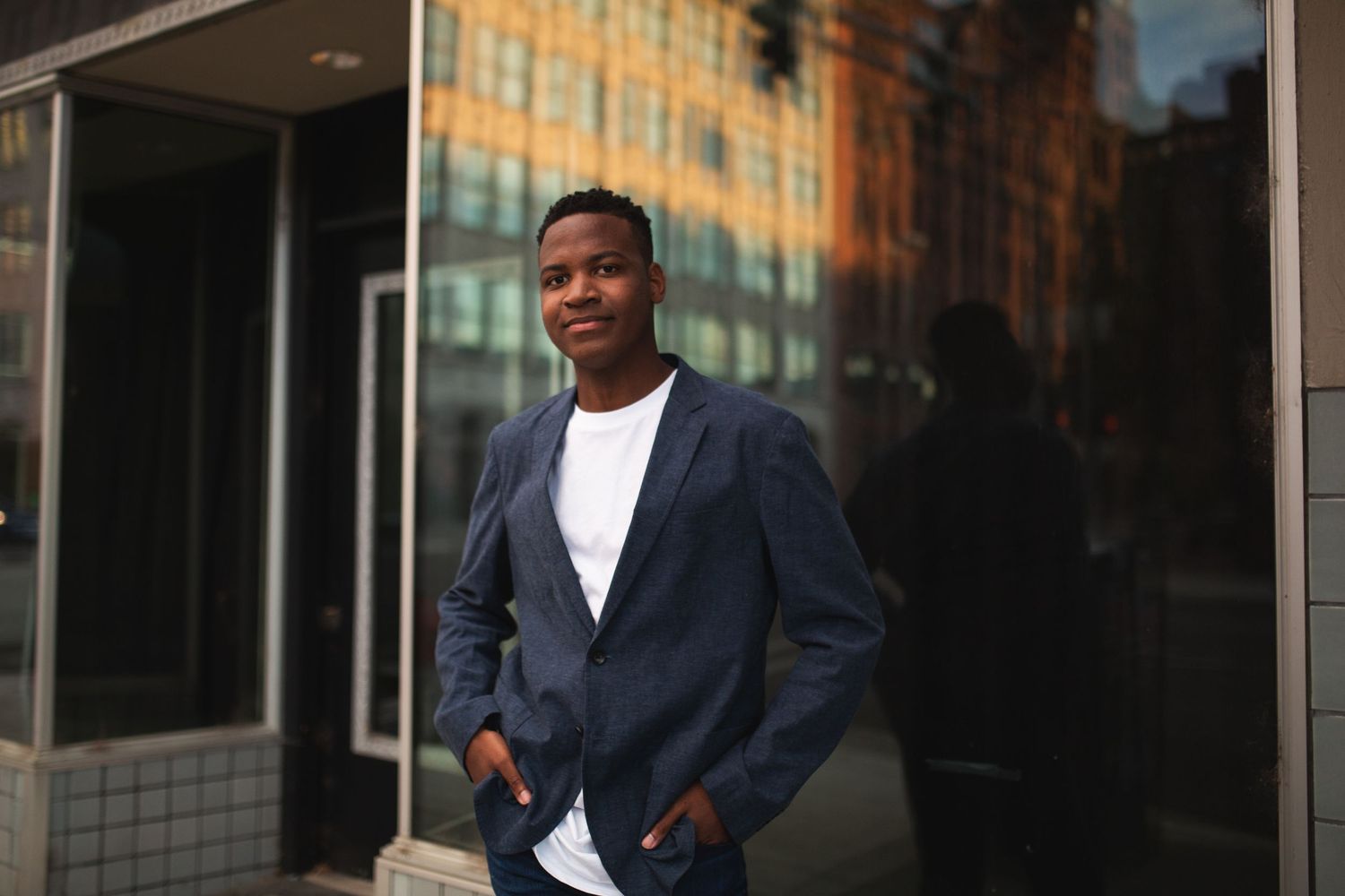 Young Black man in navy blazer and white shirt smiling outdoors near glass building in urban setting.