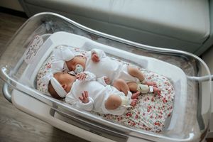 Twin newborn babies sleeping peacefully in hospital bassinet wearing white floral outfits.