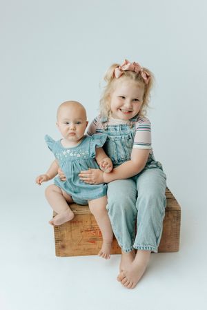 Smiling toddler girl in denim overalls sitting with baby sister in denim romper on wooden crate.