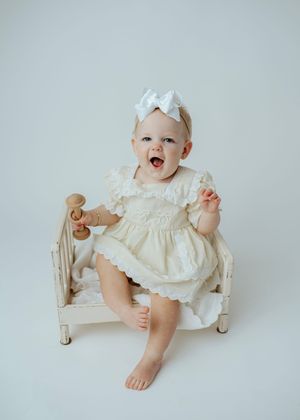 Happy baby girl in cream dress with white bow headband sitting on a miniature white bed.