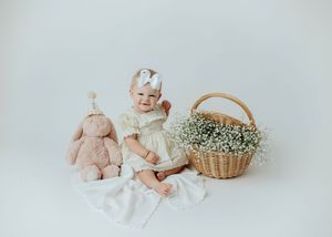 Adorable baby girl in cream dress smiling beside a pink bunny and flower basket on white.