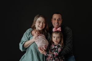 Happy family of four with newborn baby and toddler daughter posing against a dark black background.