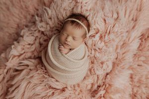 Sleeping newborn baby girl swaddled in cream knit wrap with gold headband on pink fluffy fur backdrop.