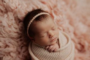 Close-up of peaceful newborn baby girl wrapped in cream knit swaddle wearing delicate pearl headband on pink fur.