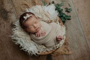Newborn baby swaddled in beige wrap with bow headband sleeping in basket with white fur and eucalyptus.