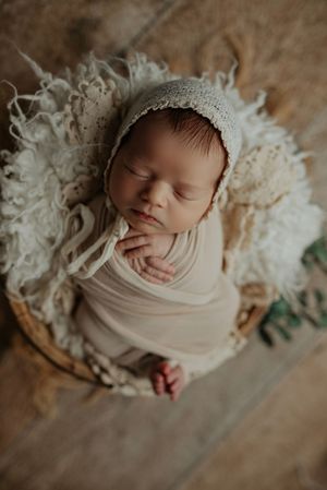 Sleeping newborn baby in cream wrap and lace bonnet posed in rustic wicker basket with white flokati fur.