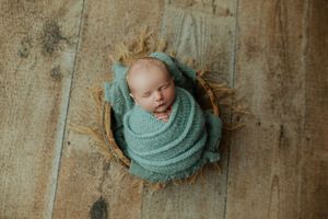 Sleeping newborn baby wrapped in teal knit swaddle nestled in wooden bowl on rustic floor.