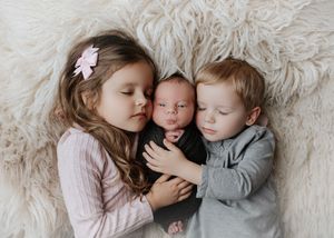 Two young siblings tenderly cuddling a sleeping swaddled newborn baby on a fluffy white rug.