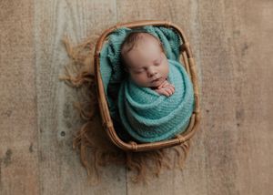 Sleeping newborn baby wrapped in teal knit blanket nestled in a small wicker basket on wood floor.