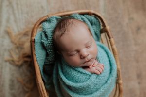 Close-up of peaceful newborn baby swaddled in teal wrap sleeping in a wicker basket.