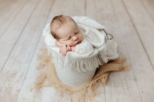 Newborn baby resting chin on hands sleeping in a white bucket with cream blanket on white floor.