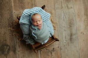 Sleeping newborn baby swaddled in teal wrap nestled in a wooden bowl on wood floor.