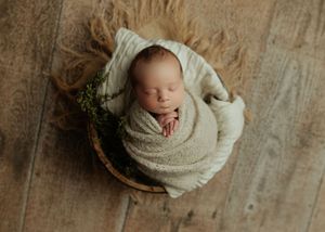 Sleeping newborn baby wrapped in cream knit blanket nestled in a wooden bowl with green foliage accents.
