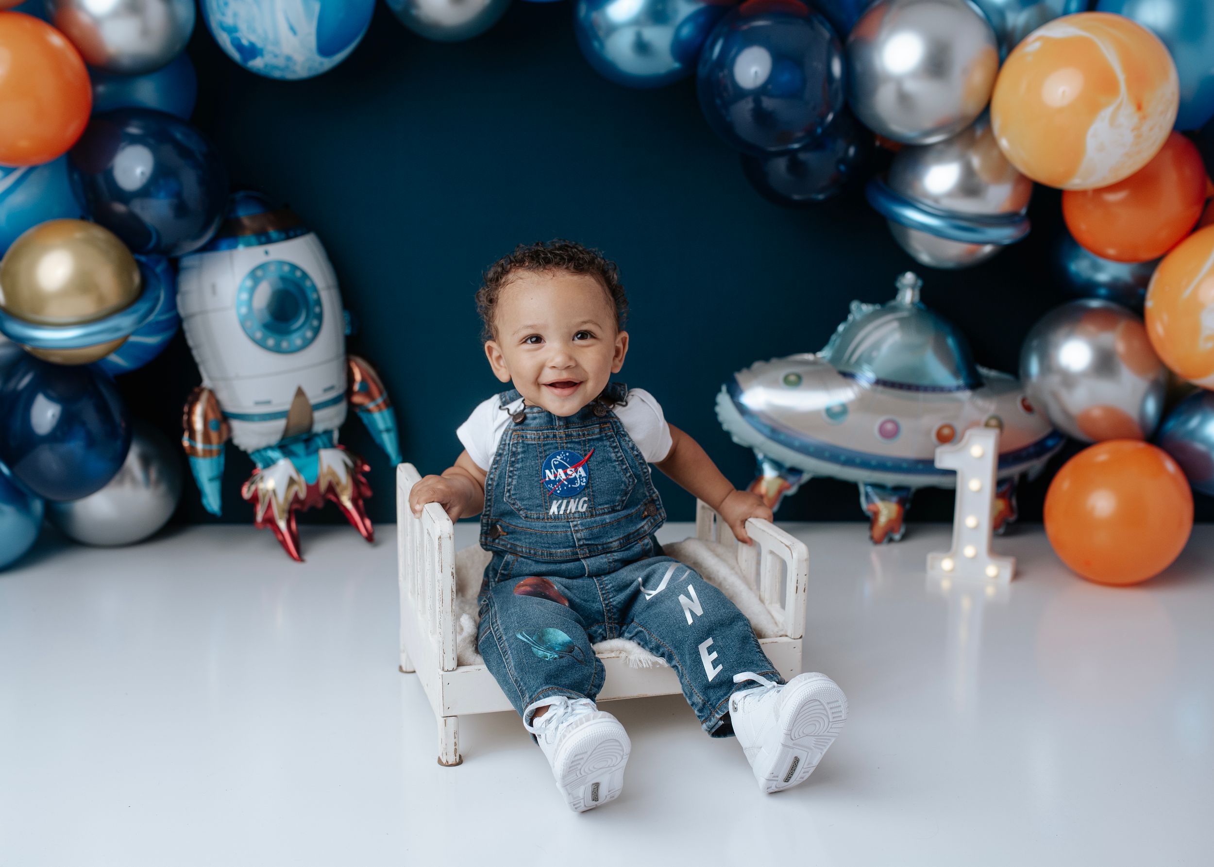 Happy toddler boy in NASA overalls laughing at a space-themed first birthday cake smash.