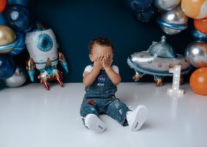 Toddler boy in NASA overalls covering his face at a colorful space-themed birthday setup.