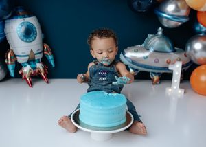 Baby boy in denim overalls smashing blue cake at space-themed first birthday party with rocket balloons.