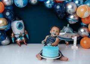 One-year-old boy enjoying blue cake smash with space theme balloons and rocket decorations behind him.